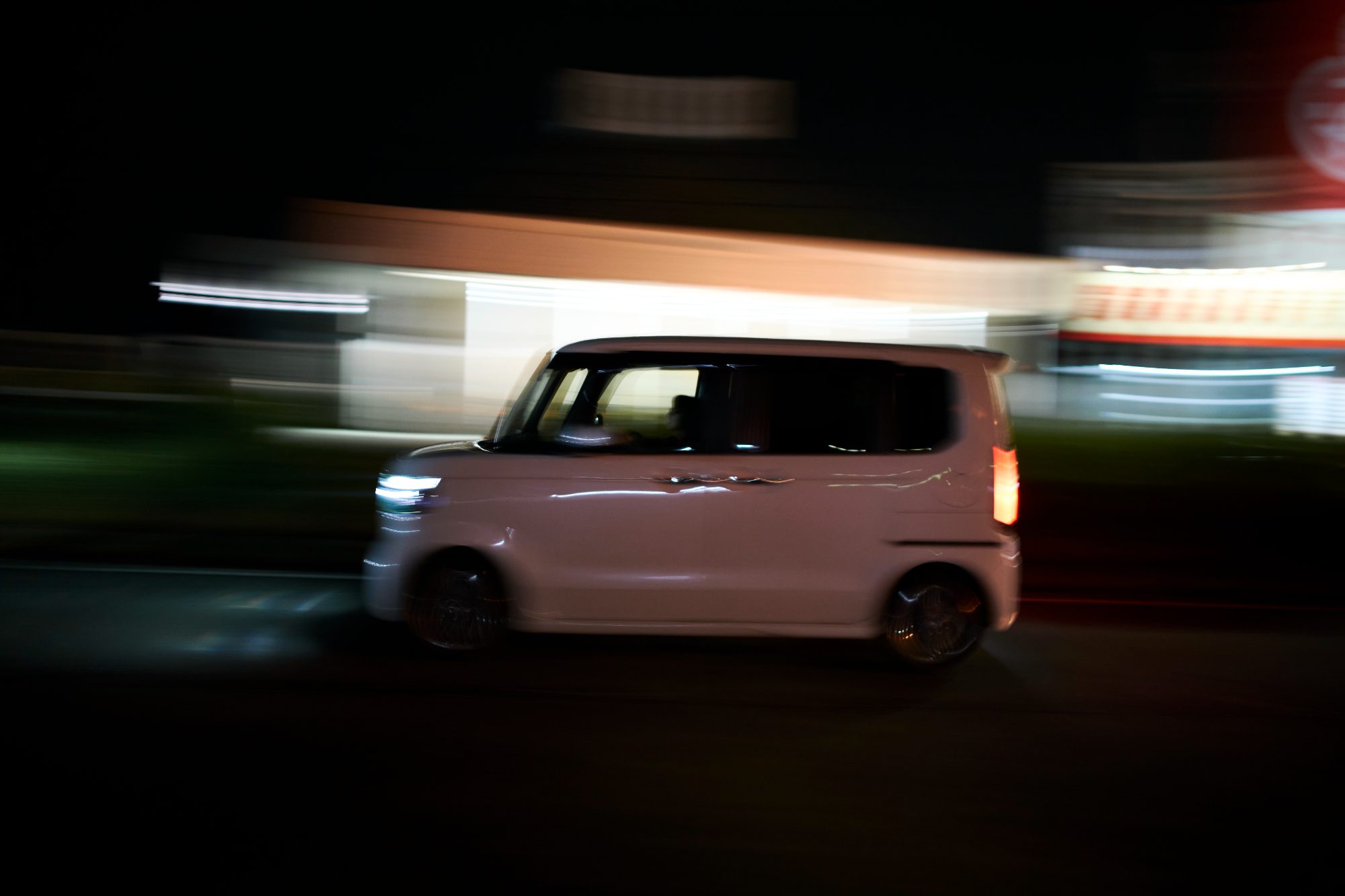 A white kei car blurred past a lit storefront at night