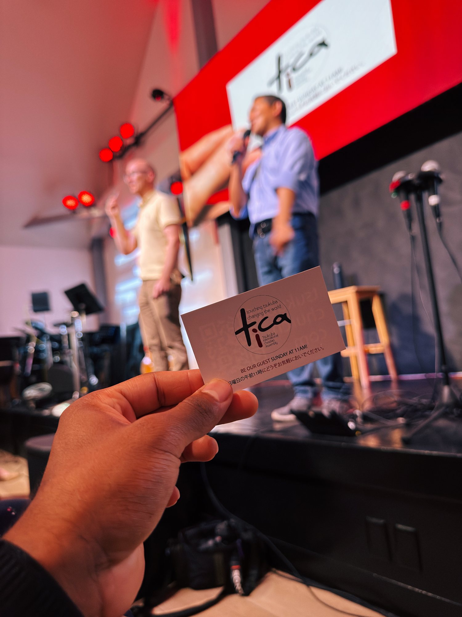 A hand holding a TICA invitation card in front of the stage during the Sunday service
